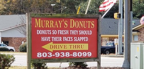 Sign for Murray's Donuts, advertising fresh donuts with a humorous tagline and drive-thru service. Phone number included.