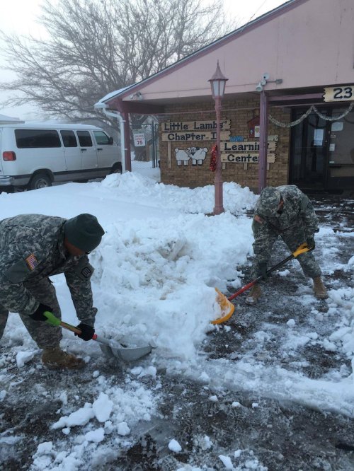 Two soldiers in military uniforms shovel snow outside a building labeled "Little Lambs Learning Center."