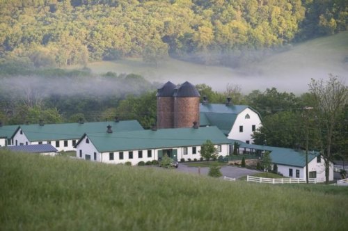 A scenic view of a white barn with green roofs, surrounded by lush hills and misty trees in the background.