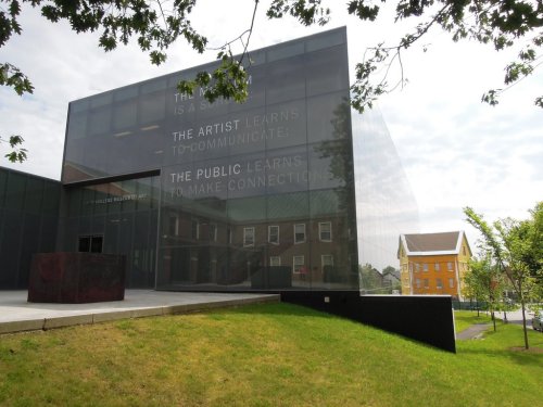 Modern building with glass facade featuring text about art and communication, surrounded by green grass and trees.