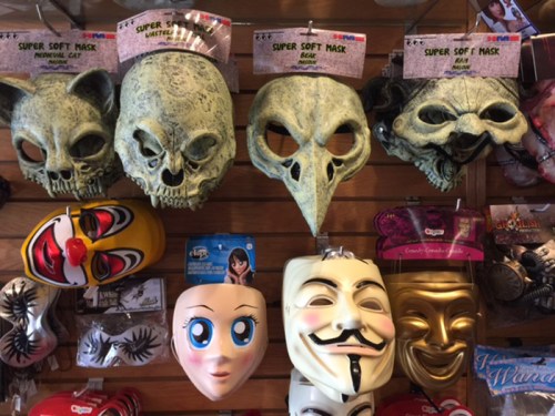 A display of various masks, including skulls, a bird, a clown, and a Guy Fawkes mask, hanging on a wall.