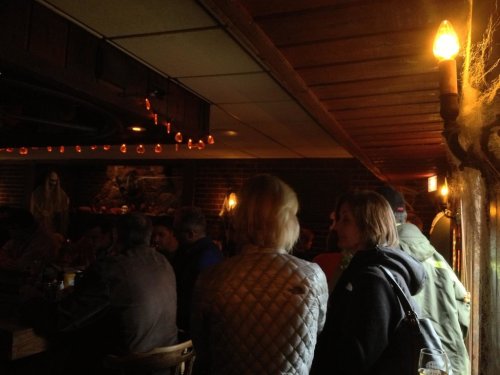 A dimly lit bar scene with patrons seated at a wooden table, chatting and enjoying drinks in a cozy atmosphere.