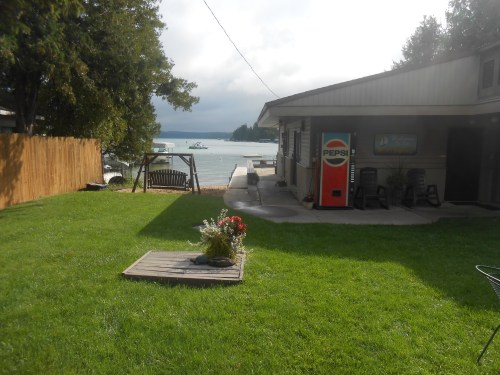 A grassy yard with a Pepsi vending machine, a swing, and a view of a lake and cloudy sky in the background.