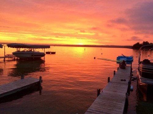 A vibrant sunset over a calm lake, with boats docked and a person sitting on a pier, reflecting warm colors on the water.