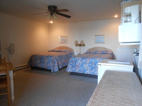 A simple hotel room with two beds, a dining table, and a small kitchenette, featuring light-colored walls and carpet.
