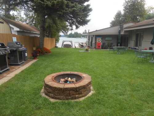 A grassy area with a fire pit, grills, and seating, overlooking a lake with trees and cabins in the background.