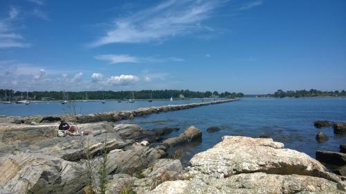 A serene coastal scene with rocky shores, a calm blue sea, and boats in the distance under a clear sky.