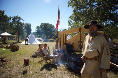 A historical reenactment scene with two men in period clothing, tents, and a campfire under a clear blue sky.