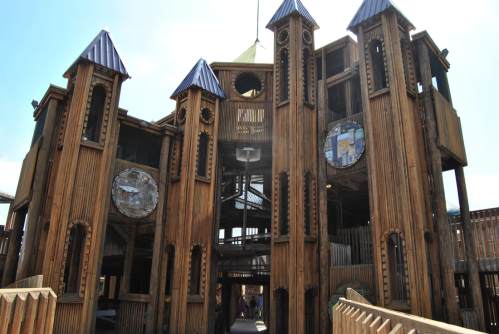A large wooden playground structure with towers and slides, featuring a whimsical design and blue roofs.
