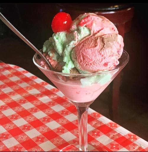 A colorful ice cream sundae in a glass, featuring pink, green, and brown scoops, topped with a cherry.