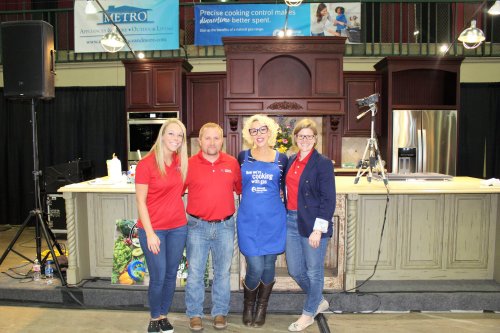Four people pose together in a kitchen setting, smiling, with a cooking demonstration backdrop.