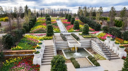 A vibrant garden with colorful flower beds, a central fountain, and neatly arranged pathways under a cloudy sky.