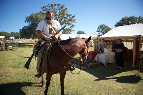 A bearded man on horseback holds a rifle, with tents and a woman in period clothing in the background.