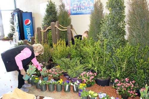 A woman arranges potted flowers and plants in a garden display, while another person works in the background.