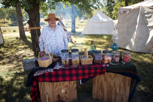 A vendor stands behind a table with jars of colorful goods, surrounded by tents and trees at an outdoor market.
