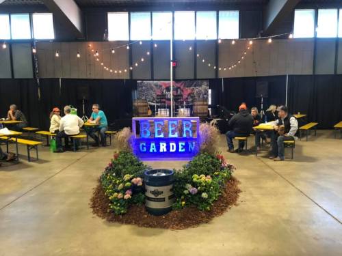 A lively beer garden with a neon sign, surrounded by flowers and seating, filled with people enjoying the atmosphere.