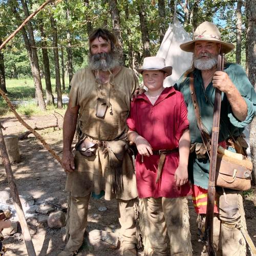 Three individuals in historical attire stand together in a wooded area, with a tent and campfire in the background.