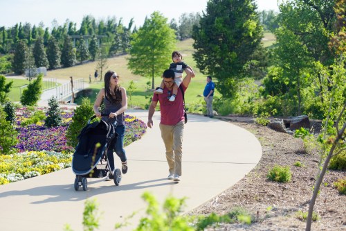A family walks through a park, with a woman pushing a stroller and a man carrying a child on his shoulder.