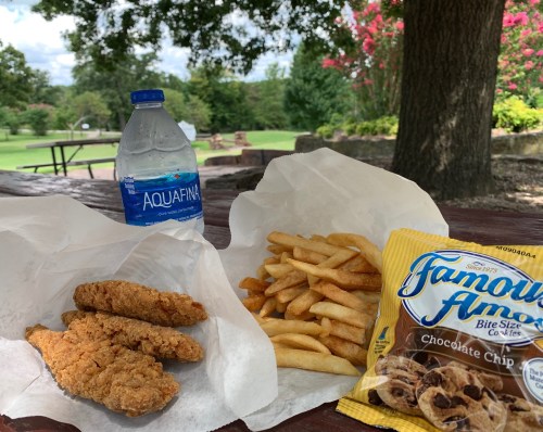 A picnic table with a bottle of water, fried chicken strips, French fries, and a bag of chocolate chip cookies.