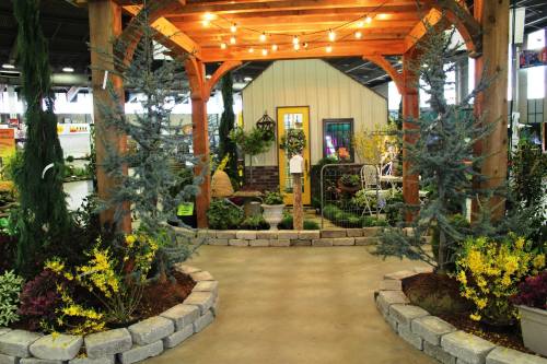 A garden display featuring a wooden pergola, plants, and a cozy seating area with decorative lights.