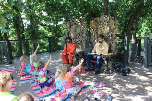 Two musicians perform on a stage in a park while children in colorful shirts sit on the ground, raising their hands.