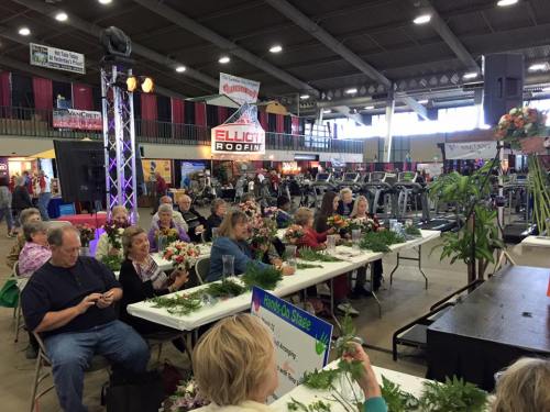 A group of people seated at tables, participating in a floral arrangement workshop at a large indoor event.