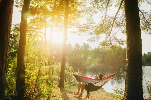 A person relaxes in a hammock between trees by a serene lake, bathed in warm sunlight.