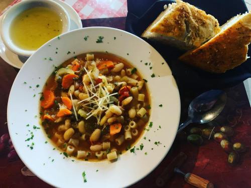 A bowl of vegetable soup with pasta, topped with cheese, alongside a slice of garlic bread and a small cup of broth.
