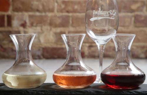 Three glass decanters with white, rosé, and red wine, alongside an empty wine glass with a logo, set against a brick background.