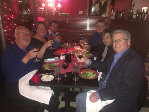 A group of six people at a restaurant, smiling and toasting with glasses of red wine over a table with food.