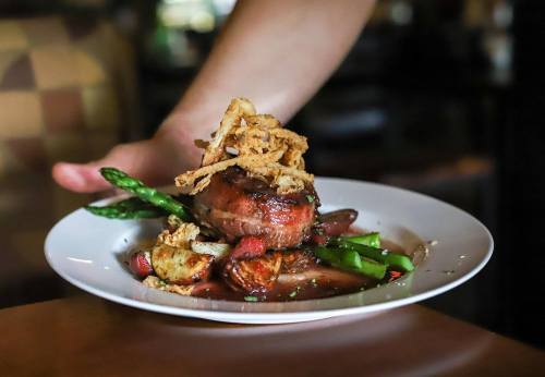 A gourmet dish featuring a meat patty topped with crispy onions, asparagus, and roasted vegetables on a white plate.