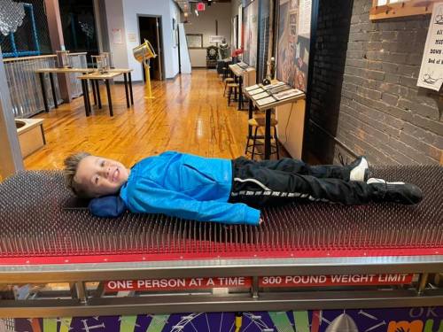A child lies on a bed of nails, smiling, in a museum exhibit with wooden floors and various displays in the background.