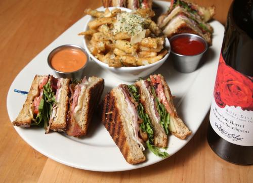 A plate with stacked sandwiches, crispy fries, and a bottle of wine, garnished with herbs and dipping sauces.