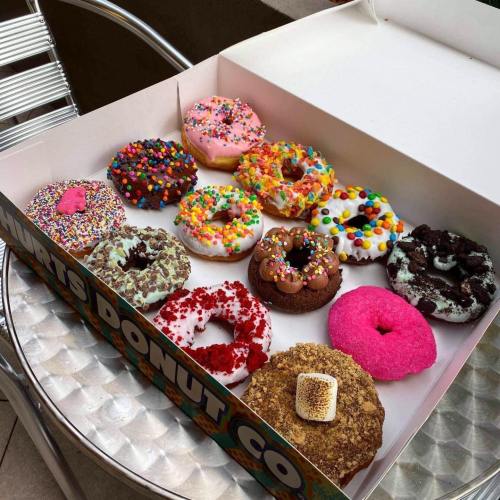 A box of colorful donuts with various toppings, including sprinkles, frosting, and chocolate, arranged on a silver table.