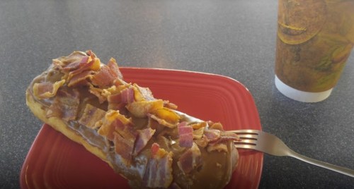 A glazed donut topped with bacon bits on a red plate, next to a coffee cup on a dark table.