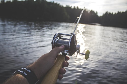 A hand holding a fishing rod with a reel, casting over a shimmering lake at sunset.