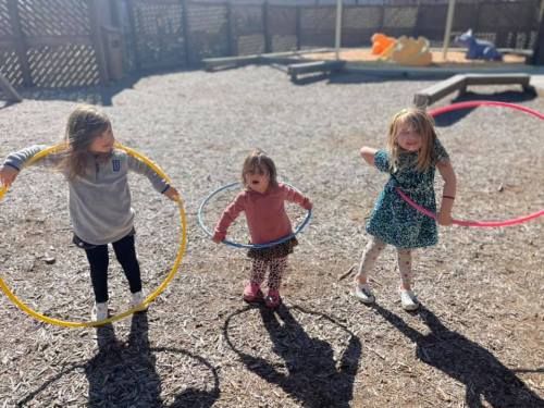 Three children play with hula hoops in a sunny playground, surrounded by wood chips and colorful play equipment.