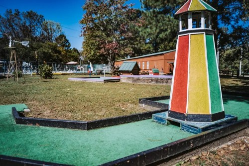 Colorful lighthouse structure on a mini-golf course, surrounded by green grass and trees under a clear blue sky.