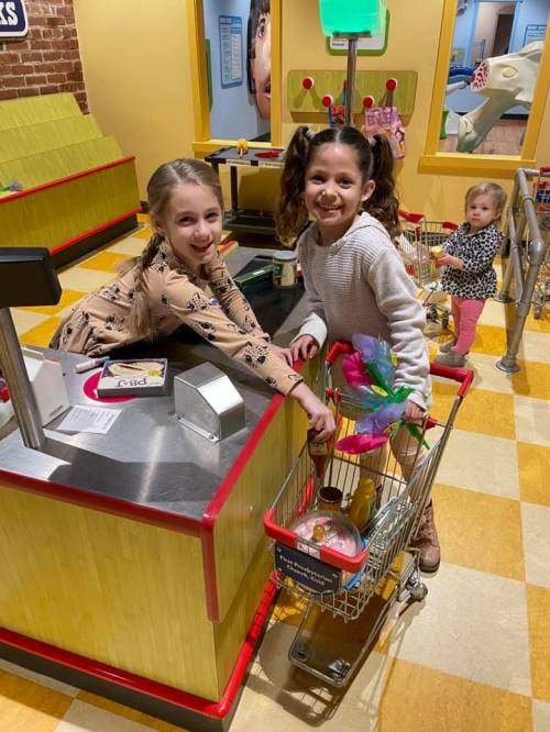 Two girls smile while pushing a toy shopping cart at a colorful play area, with a younger child in the background.