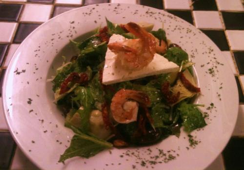A fresh salad with greens, sun-dried tomatoes, shrimp, and a wedge of feta cheese, served on a white plate.