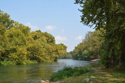 A serene river flows through a lush green landscape, surrounded by trees under a clear blue sky.