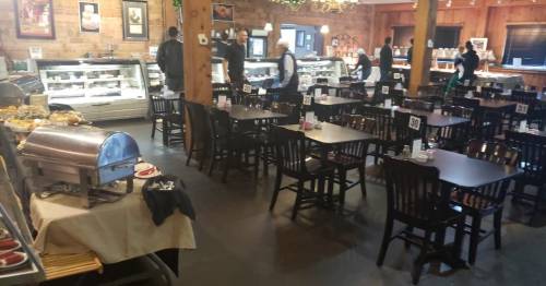 A spacious restaurant interior with wooden tables, chairs, and a buffet setup, featuring staff and patrons.