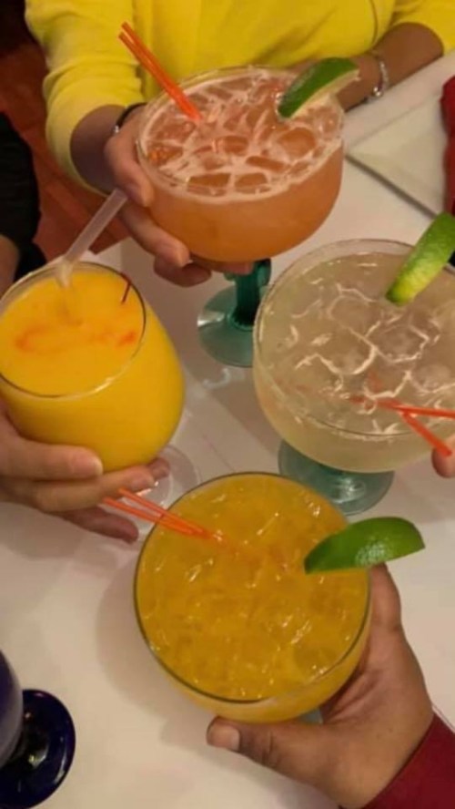 Four colorful cocktails with straws, held by hands, on a table. Drinks include orange, yellow, and clear beverages.