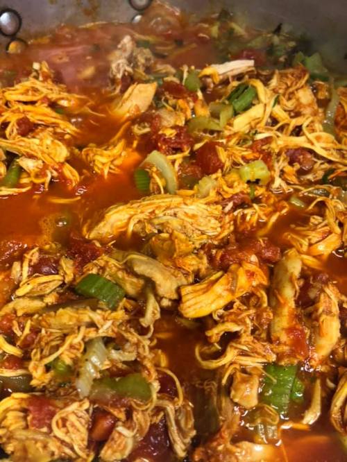 A close-up of shredded chicken simmering in a rich tomato sauce with celery and spices in a large pot.
