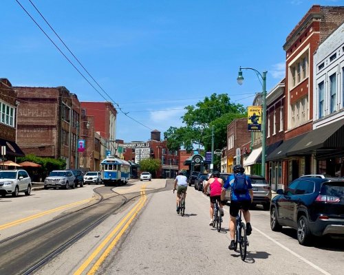 Three cyclists ride down a sunny street lined with shops and parked cars, with a vintage streetcar in the background.