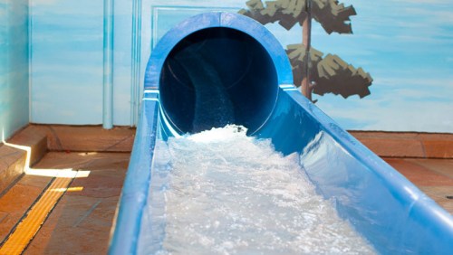 A blue water slide with flowing water, set against a painted backdrop of trees and a blue sky.