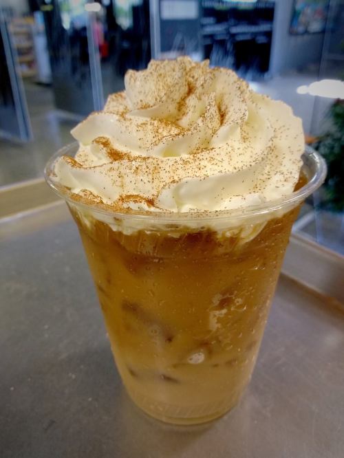 A clear cup filled with iced coffee topped with whipped cream and a sprinkle of cinnamon, placed on a tray.