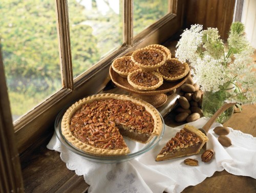 A pecan pie on a windowsill with a slice cut out, surrounded by mini pecan tarts and fresh flowers.