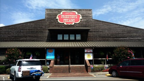 Exterior of Priester's Pecans store, featuring a wooden facade and signage, with cars parked in front on a sunny day.