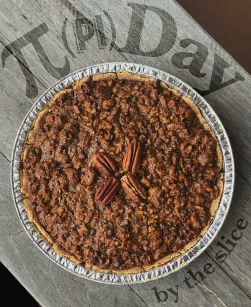 A pecan pie topped with whole pecans, displayed on a wooden surface with "Pi Day" text in the background.